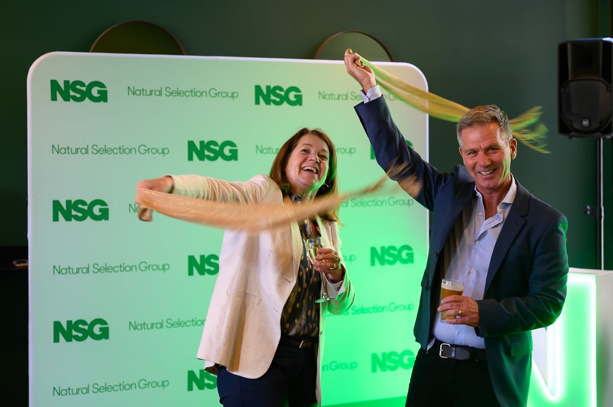 Two people smiling and holding drinks in front of a green and white Natural Selection Group branded backdrop.