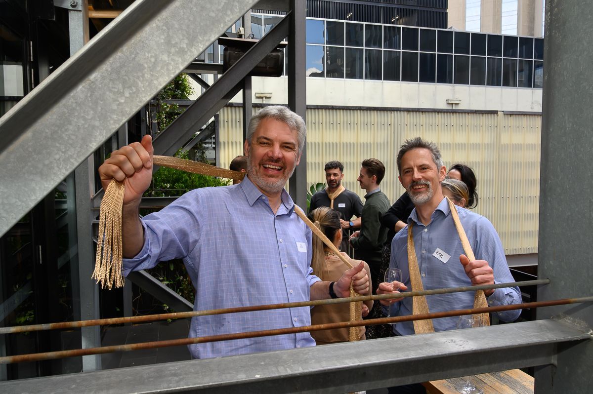 Two individuals in light-colored, long-sleeved shirts standing on a balcony, holding fringed decorative fabric.