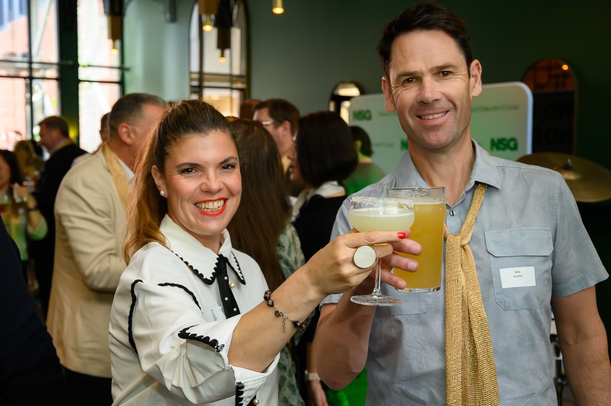 A woman and a man in a professional setting toast with drinks and smile at the camera.