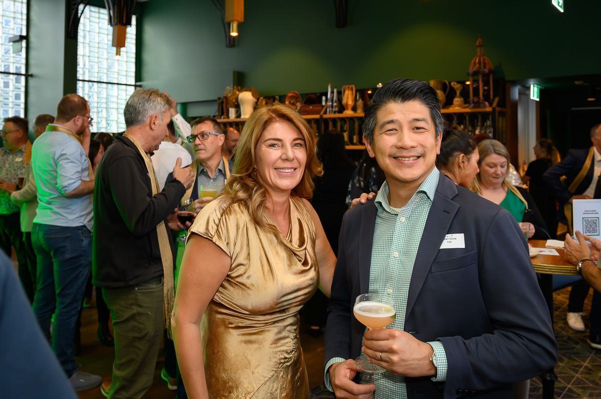 A man in a navy blazer and woman in a gold dress smile while holding drinks at a crowded, dimly lit indoor event.