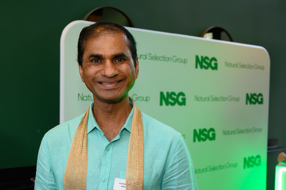 A smiling person in a light green shirt and gold scarf stands before a backdrop branded with 