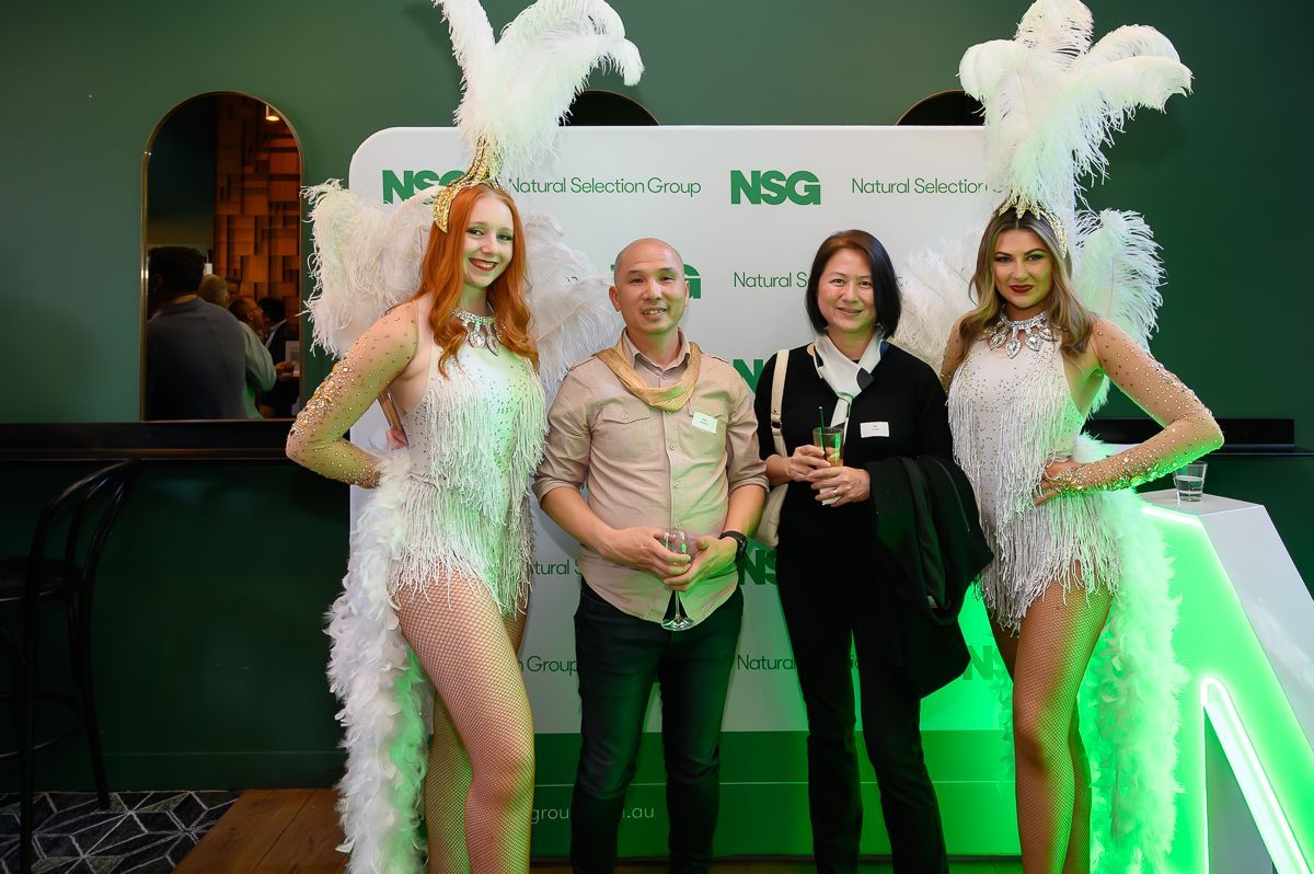 Two showgirls in white feathered costumes pose with a man and a woman in front of a green branded backdrop.