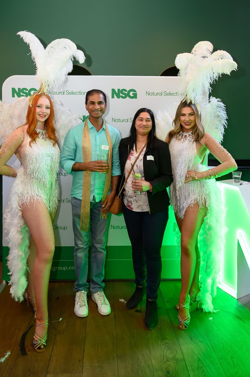 Two people stand smiling between two performers in elaborate white feathered costumes in front of an NSG-branded backdrop.