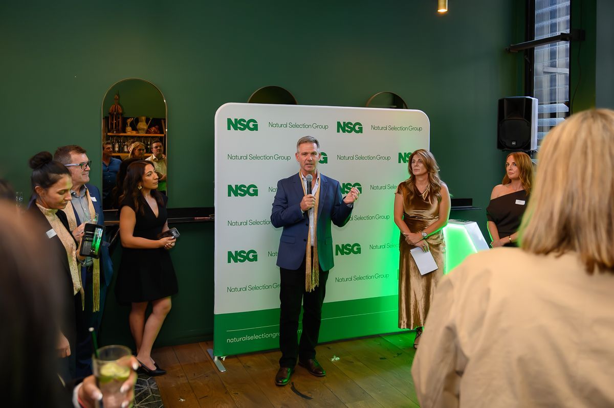 A man in a blue blazer speaks into a microphone in front of an NSG-branded backdrop while others stand nearby.