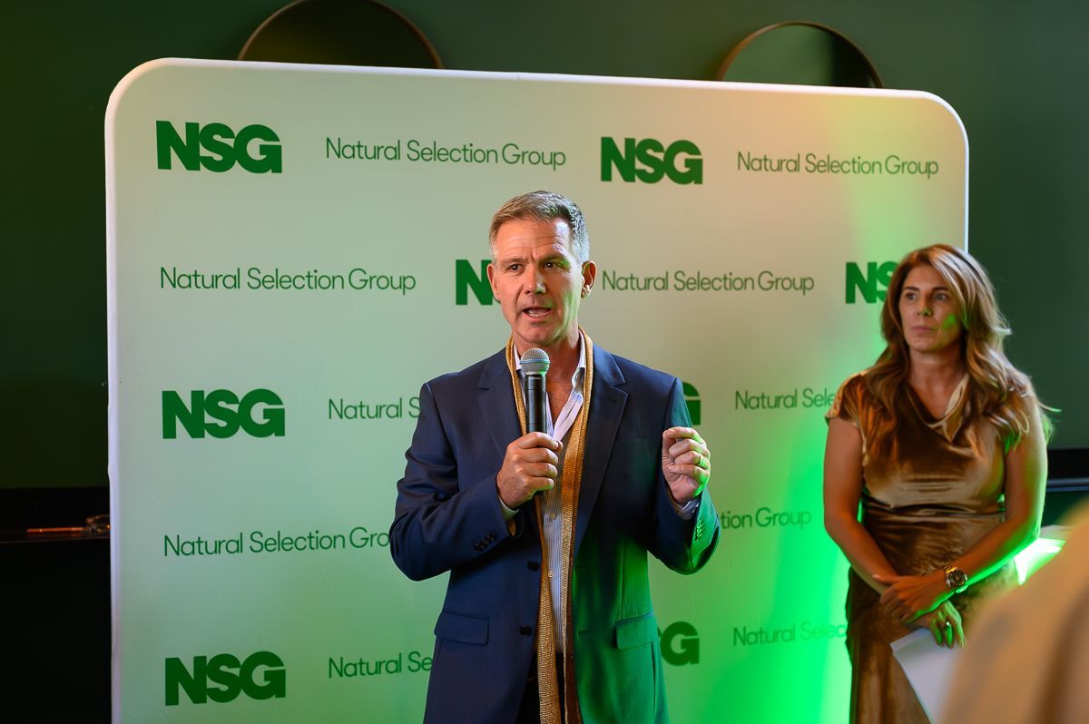 A person speaks into a microphone in front of a white branded banner, with another person standing nearby.