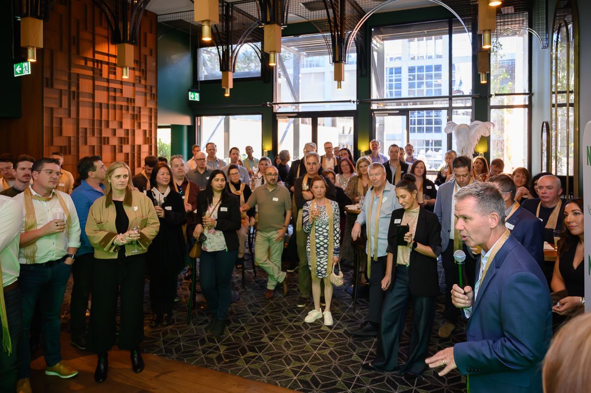 A group of people listens to a man speaking at an indoor event, with the speaker holding a microphone in the foreground.