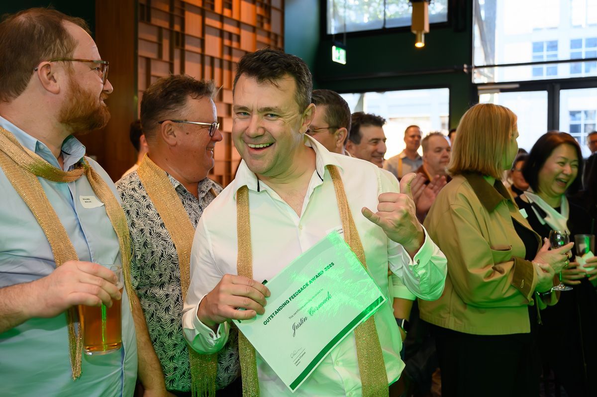 A smiling man holds a green certificate while others stand nearby at a social event with drinks and gold sashes.