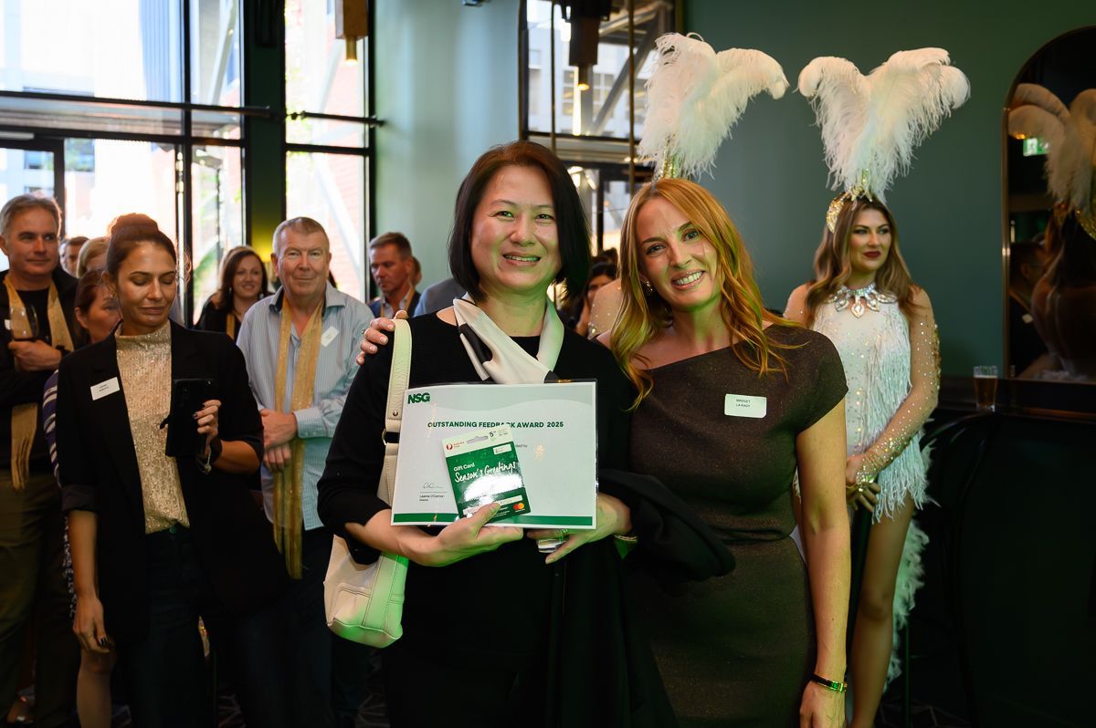 Two women smile while holding a prize certificate at an indoor event, with others and performers in the background.