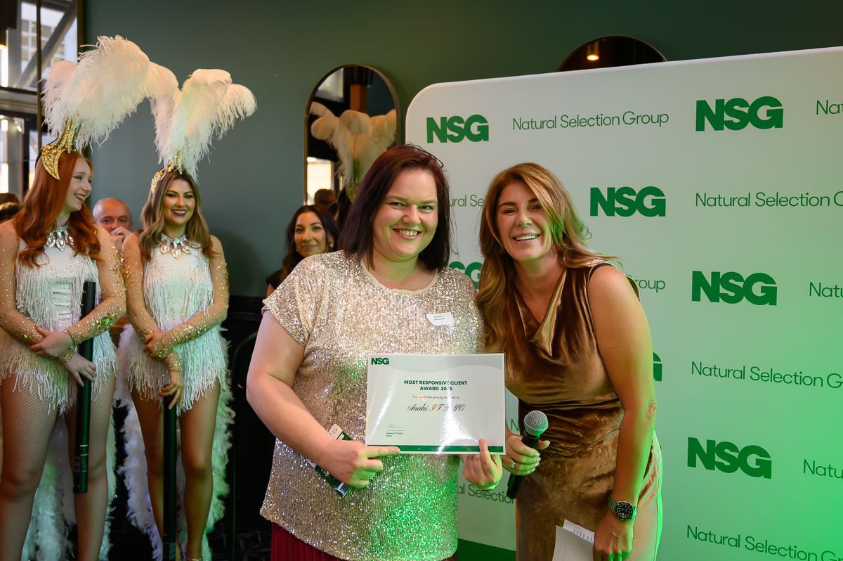 Two women stand smiling, holding a certificate in front of a green and white 