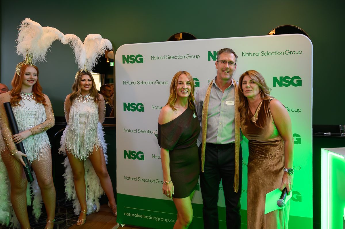 Two people stand with a man between them in front of an NSG branded backdrop, flanked by two performers in feather attire.