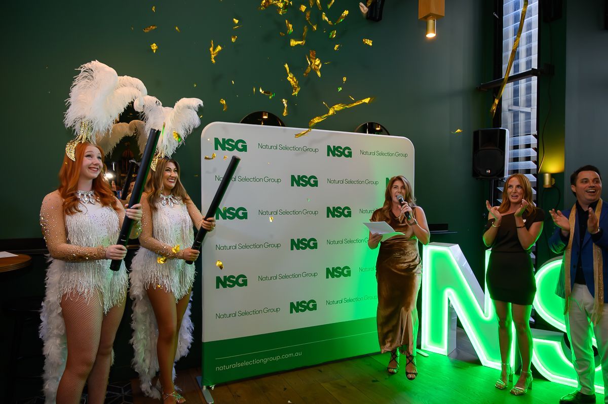 Two performers in feathered costumes and three people pose in front of an NSG-branded wall with falling gold confetti.