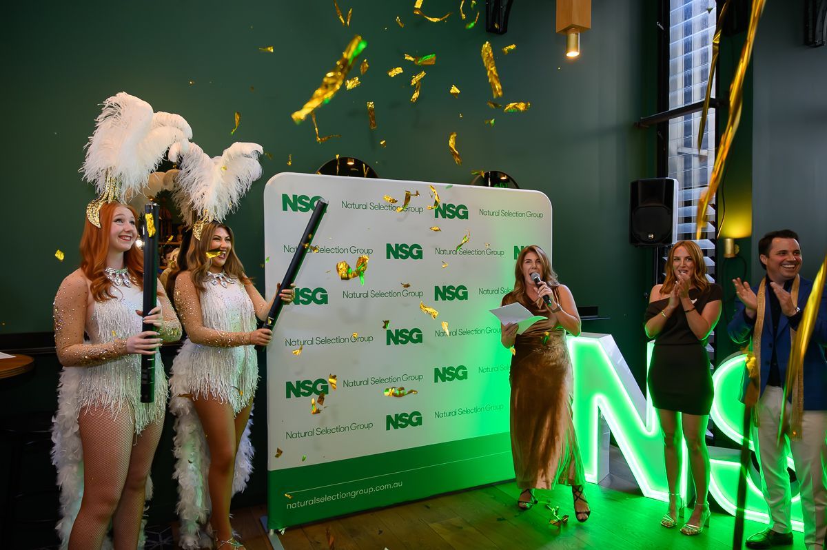 Two performers in white feathered costumes and a presenter celebrate at an event with gold confetti and an NSG sign.