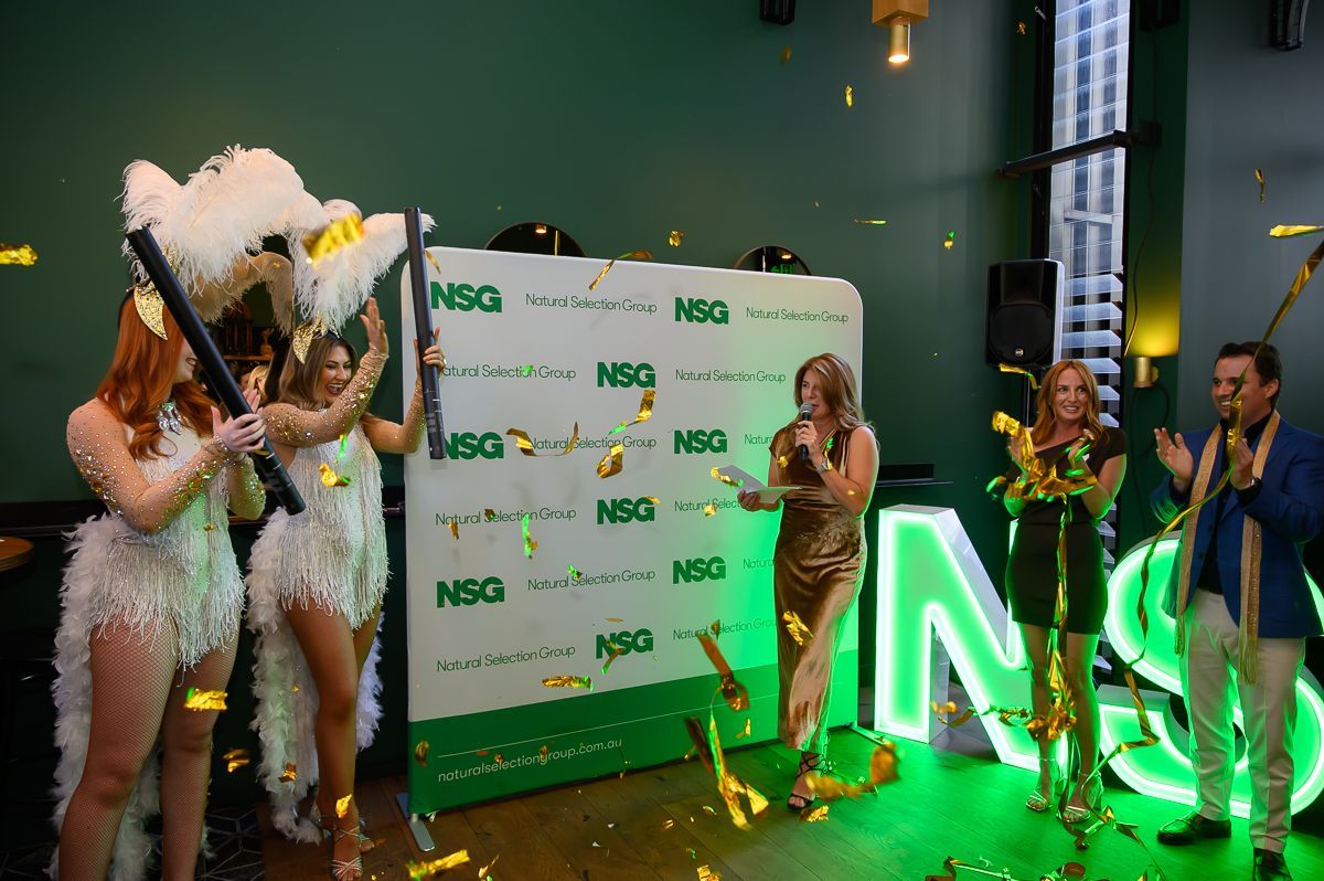 Two performers in feathered outfits and two people in formal wear celebrate in front of an NSQ branded backdrop.