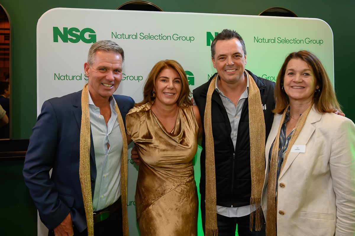 Four people smile while standing in front of a white Natural Selection Group (NSG) banner at an indoor event.
