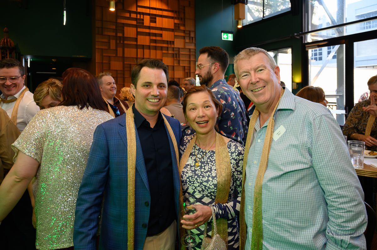 A group of people wearing gold sashes smiles at a social gathering in an indoor venue with a patterned wooden wall.