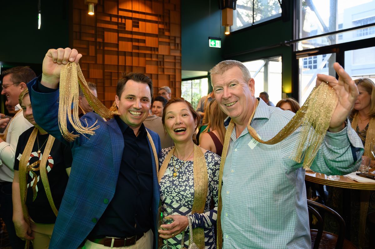Three people smiling and holding up long, gold, rope-like decorative chains at an indoor event.