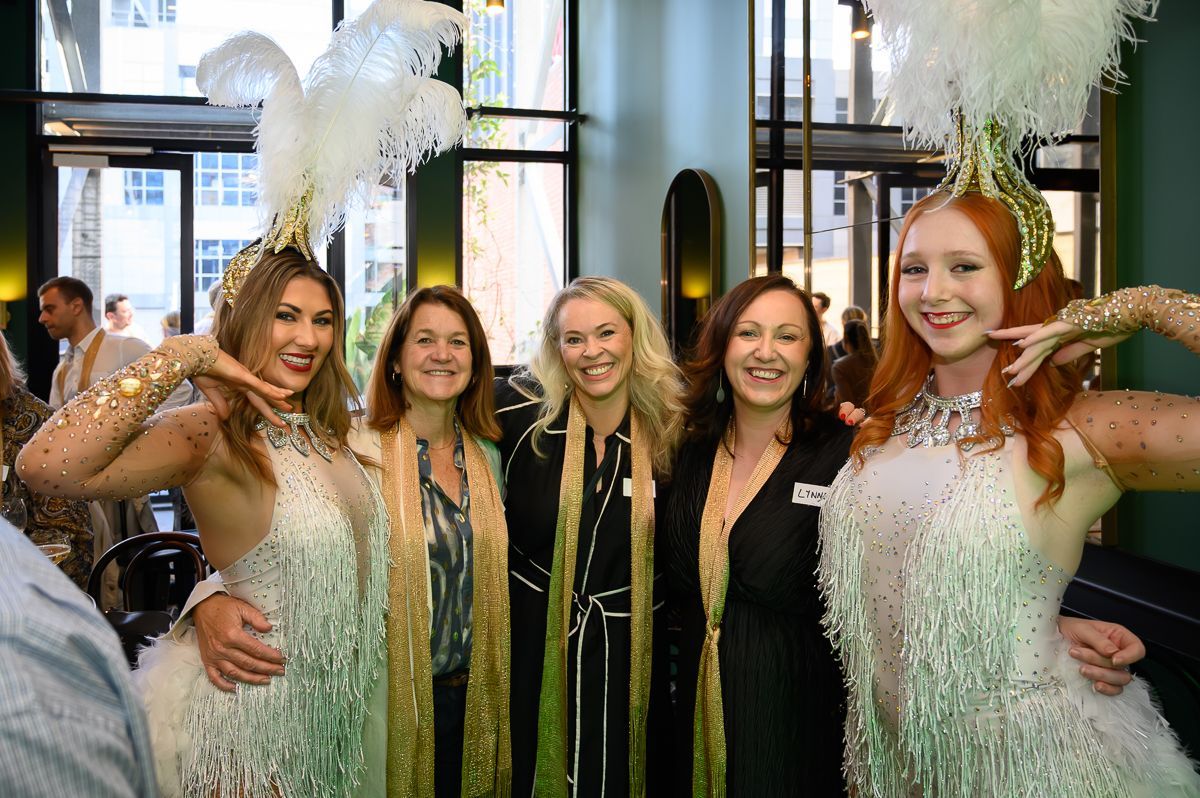 Two performers in feathered showgirl costumes stand on either side of three people smiling for a photo indoors.