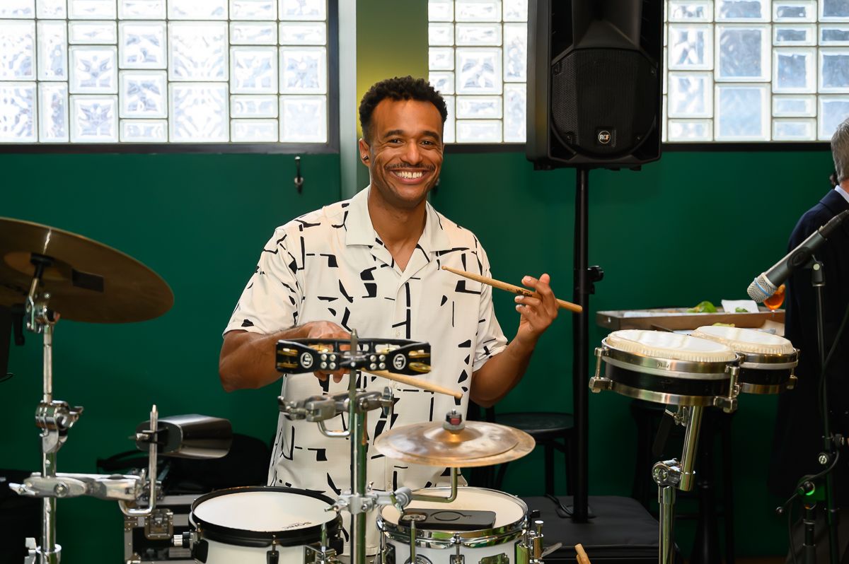 A musician smiling while playing percussion instruments, including drums and cymbals, in an indoor setting.