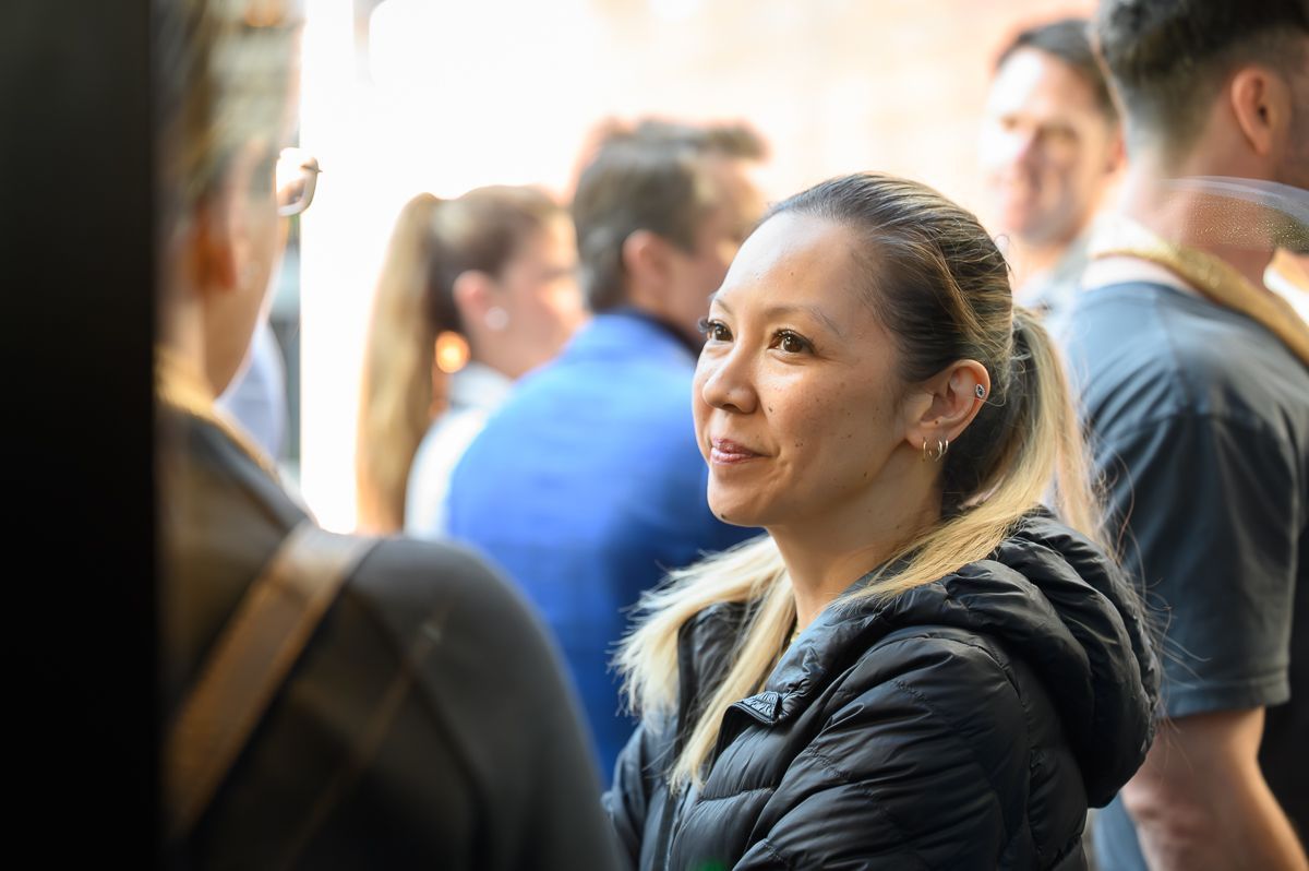 A person in a black puffer jacket looks attentively at another person, partially visible, in a crowded indoor setting.