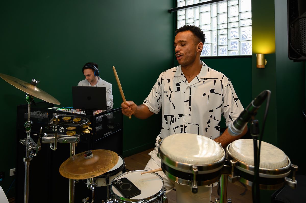 A percussionist plays bongo drums with a drumstick in a room with green walls, while another person works at a laptop.