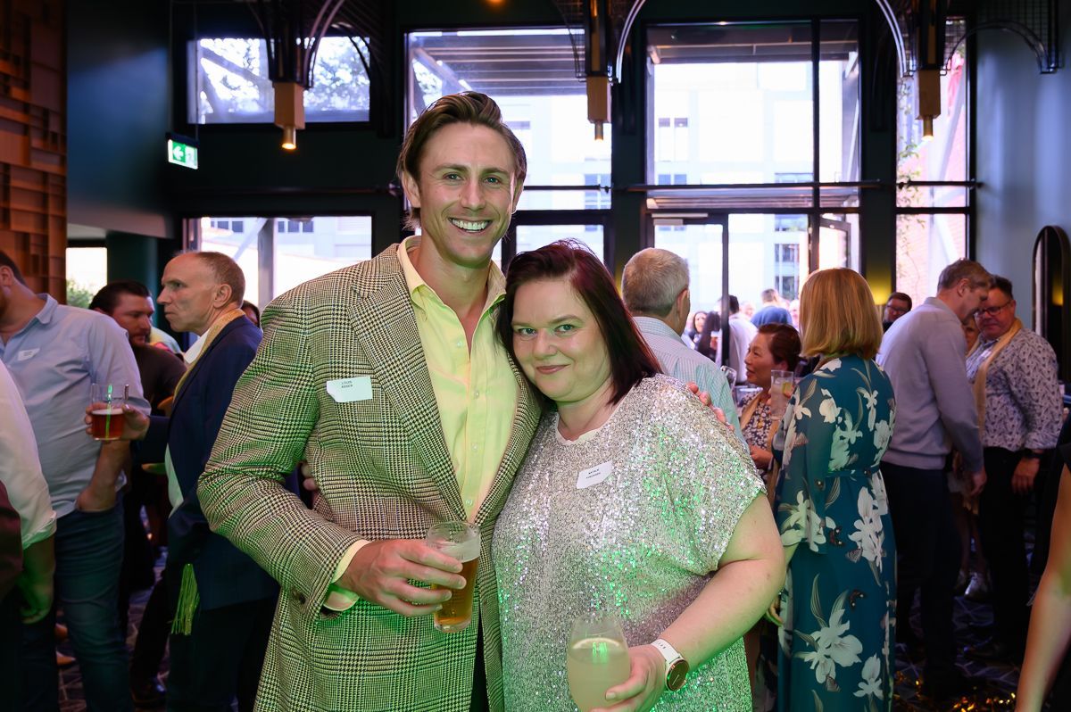 A person in a patterned blazer and another in a sequined top pose together with drinks at an indoor social gathering.