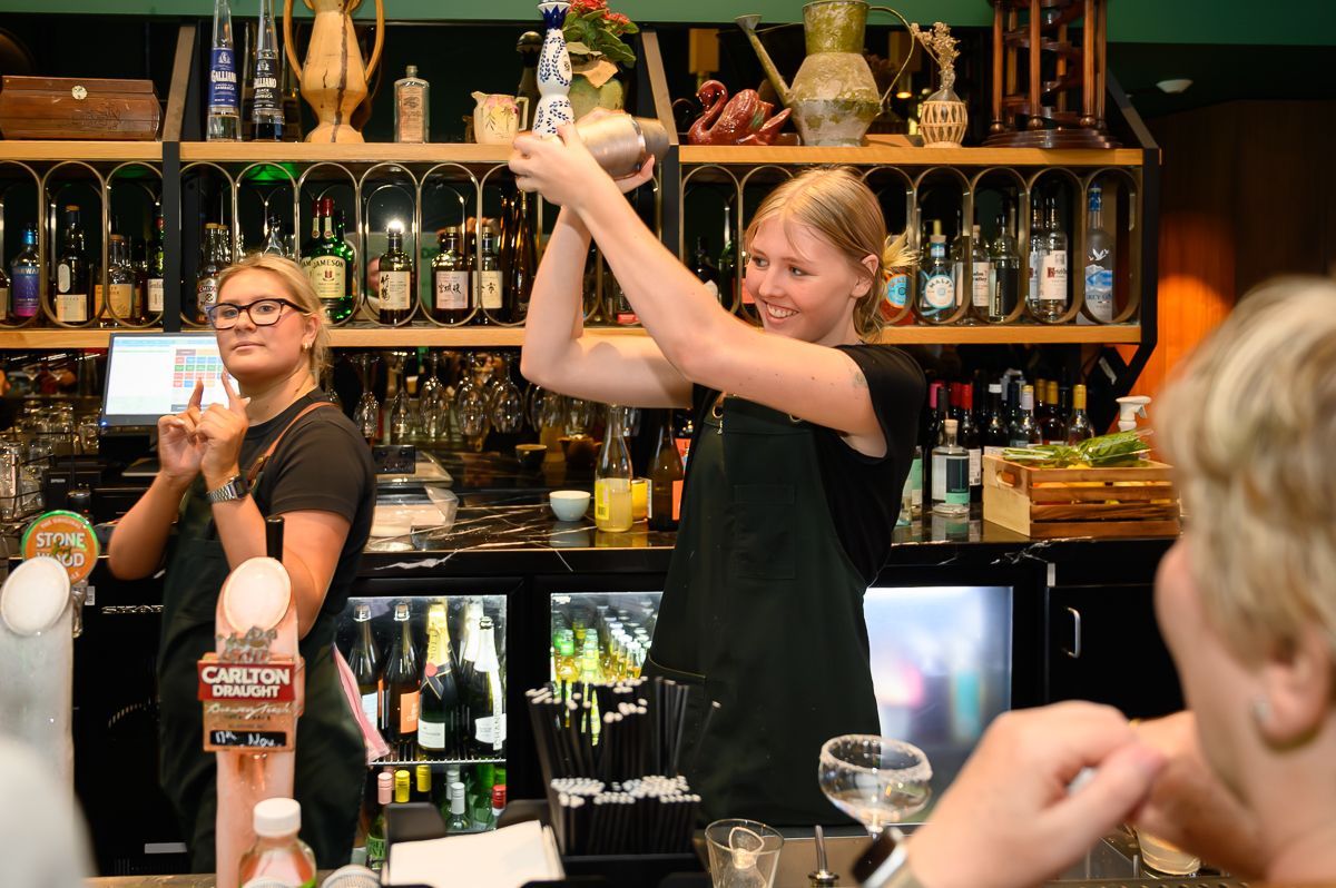 Two bartenders behind a bar counter, one shaking a cocktail, with a customer visible in the foreground.