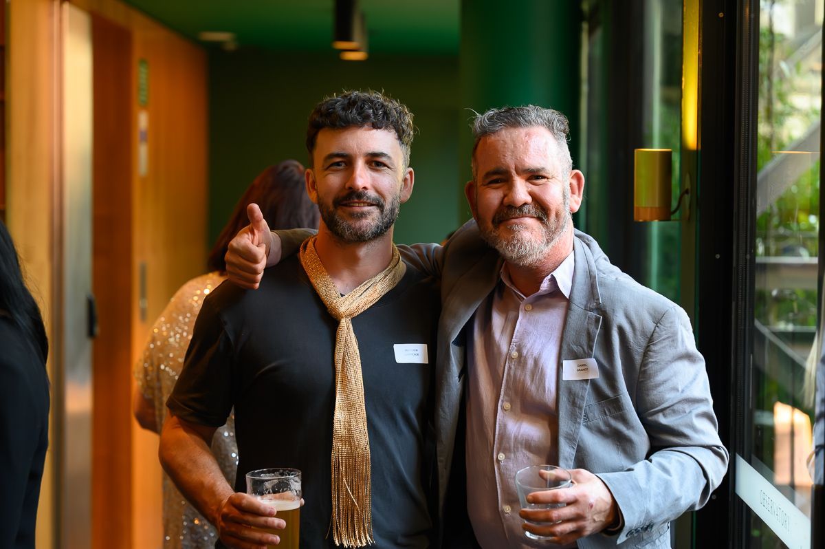 Two men smiling and posing for a photo indoors; one wears a gold sequined scarf and the other a grey blazer.