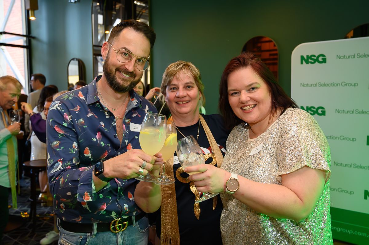 Three people smiling and raising glasses for a toast at an indoor event, standing in front of an NSG brand backdrop.