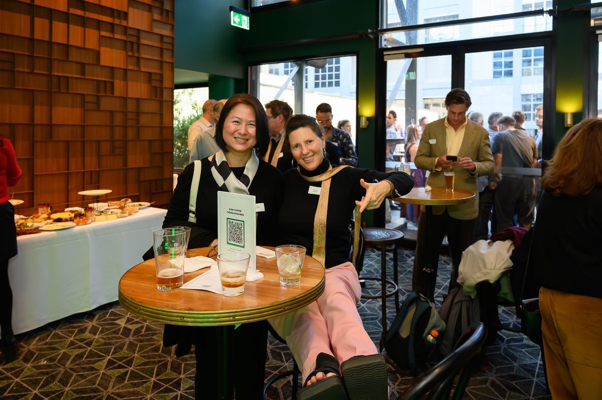 Two people smiling and posing at a small round table during an indoor social event with other guests in the background.