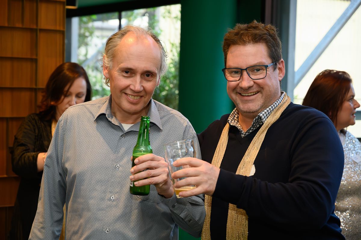 Two men smiling and holding drinks in a social setting, with two women visible in the background.