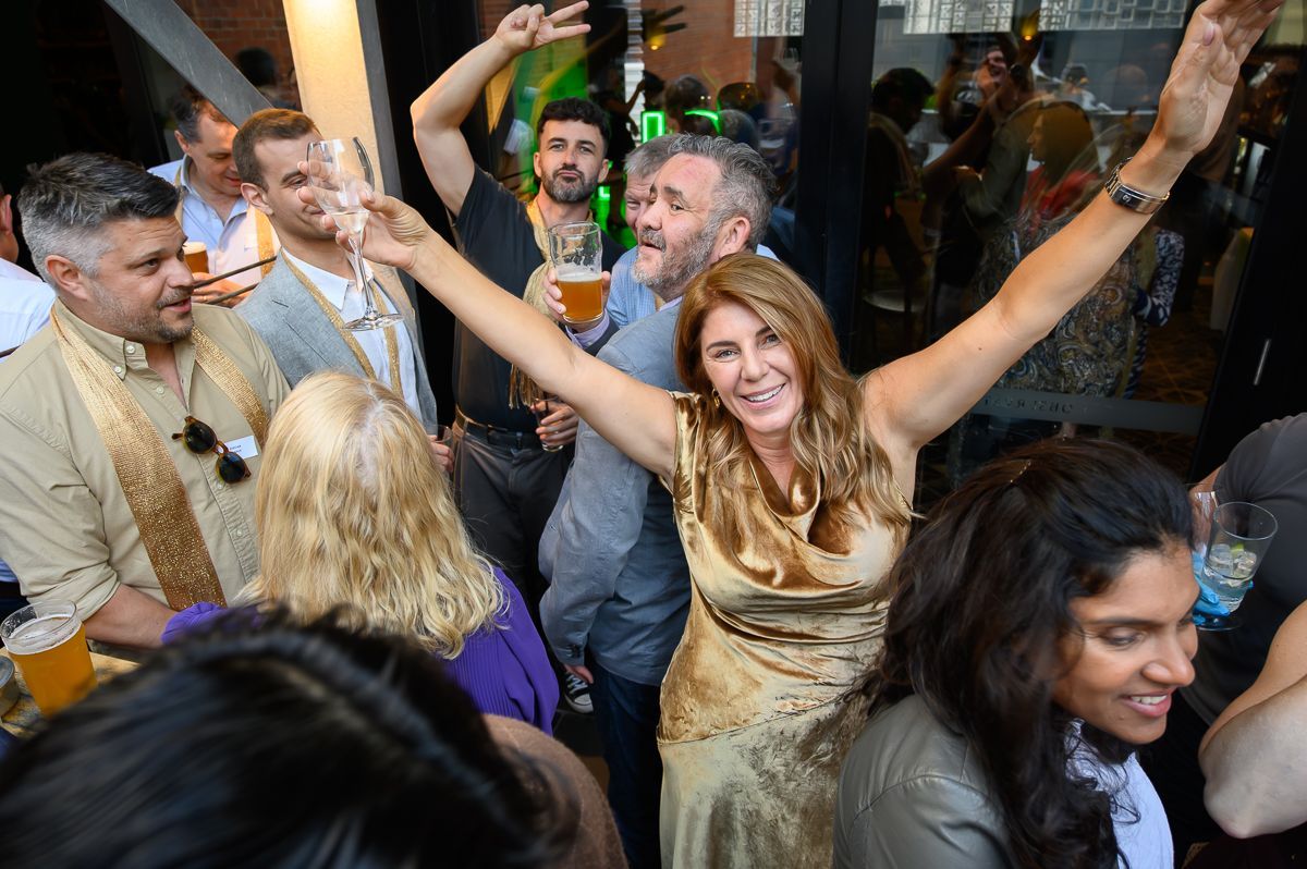 A lively group of people at an indoor event, with a smiling person in a gold dress reaching out with arms raised.