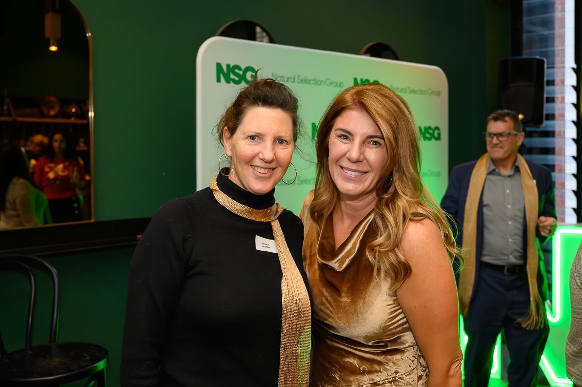 Two people smile at a corporate event with a green wall backdrop featuring the NSG logo, while a third person stands behind.