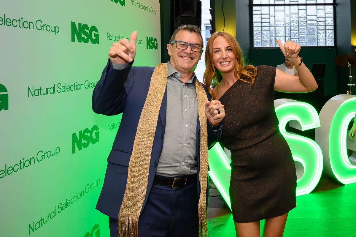 A man in a blue suit with a gold scarf and a woman in a brown dress posing together at an NSG branded event.