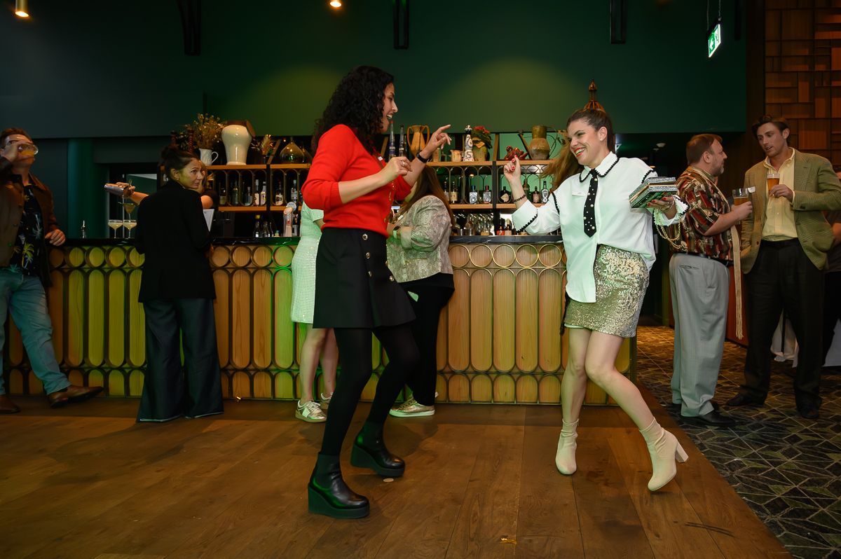 Two people dance on a wooden floor in a bar, while others stand and socialize in the background.
