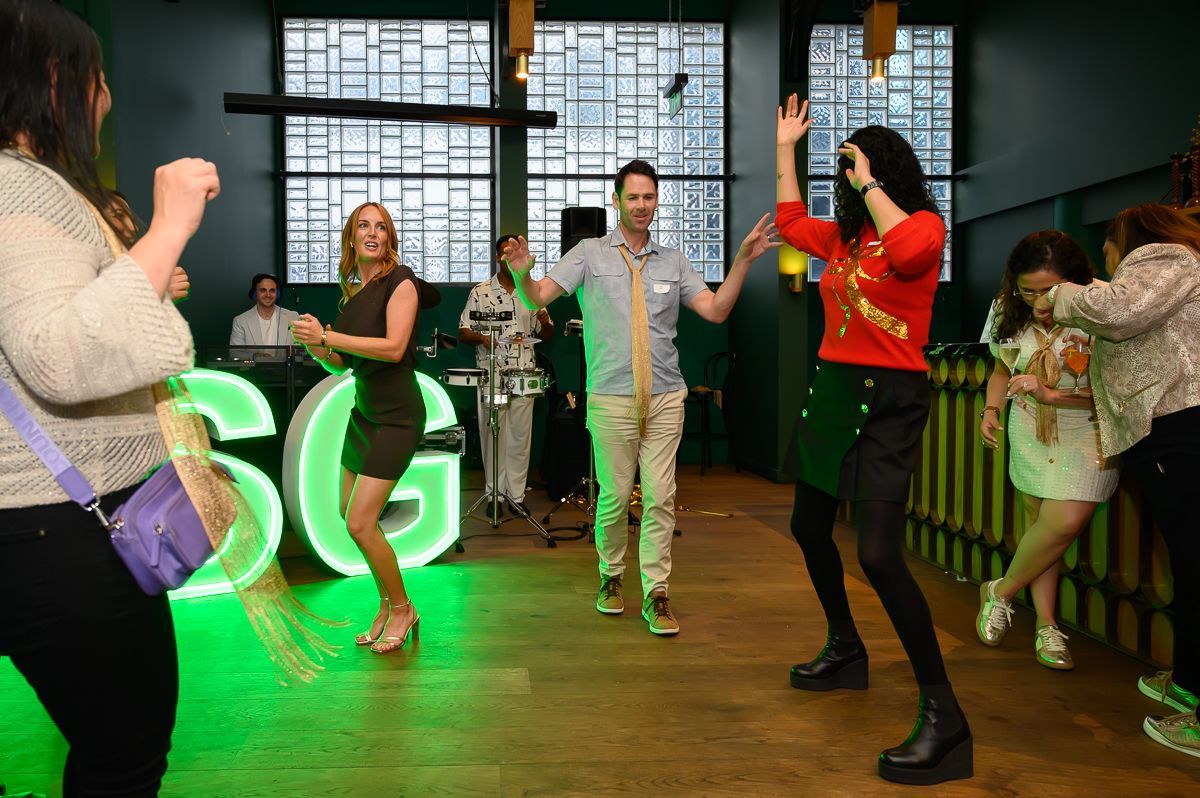 A group of people dancing in a venue with large, glowing green letters and wood floors.