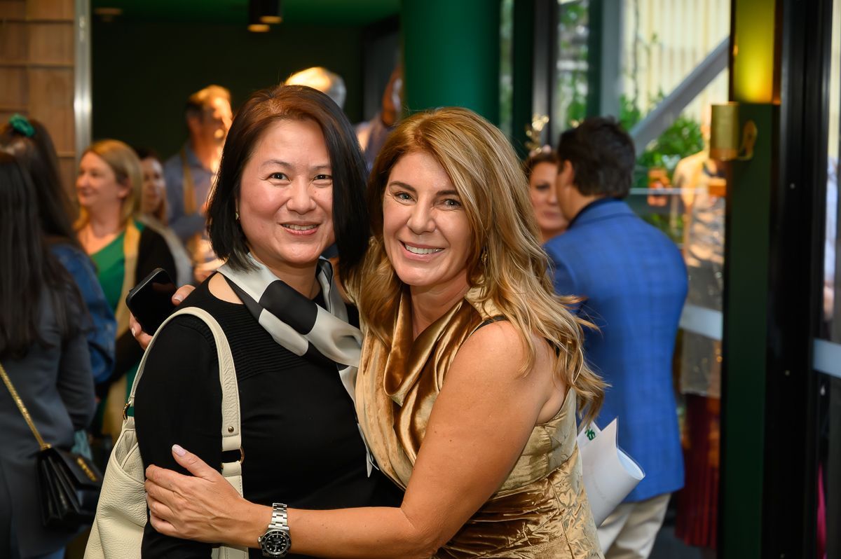 Two people embrace and smile for a photo at an indoor event, with other attendees visible in the blurred background.