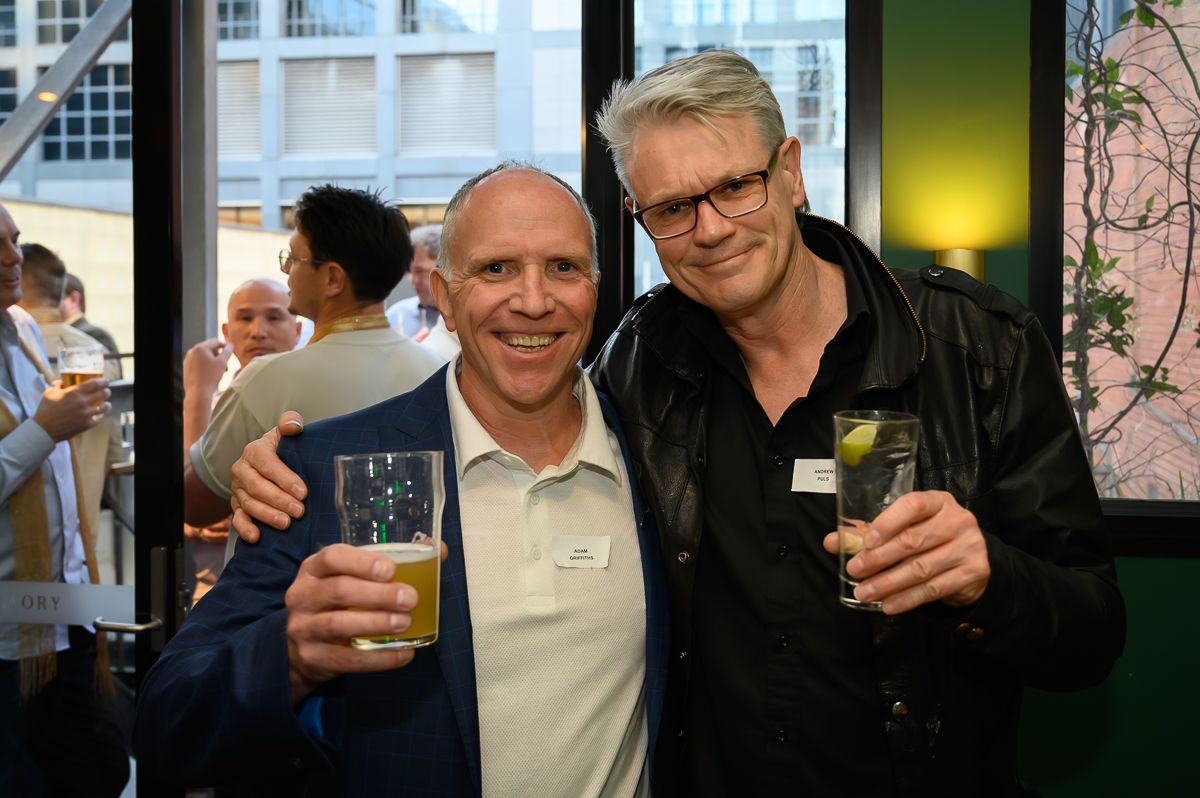 Two people stand smiling at an indoor event, both holding glasses with drinks, with others visible in the background.