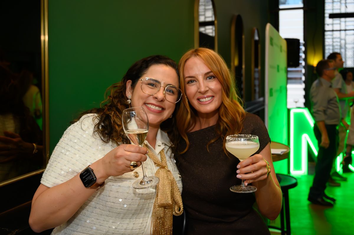 Two smiling people toast with cocktails at a social event with green lighting and a branded backdrop.