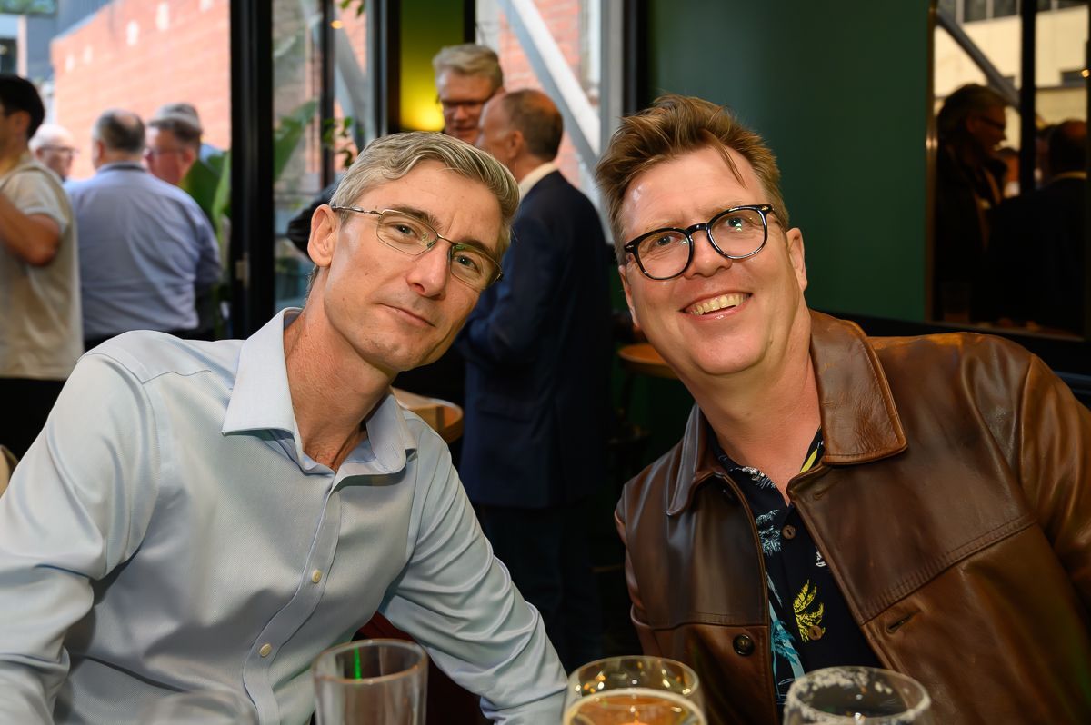 Two people sitting at a table in a dimly lit bar, one smiling and wearing a leather jacket, the other looking at camera.