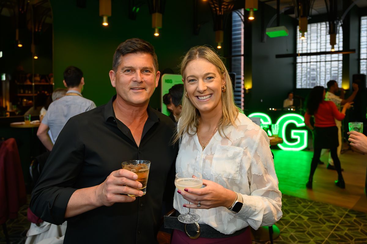 A man and woman holding drinks pose for a photo at an event with a glowing green neon sign in the background.