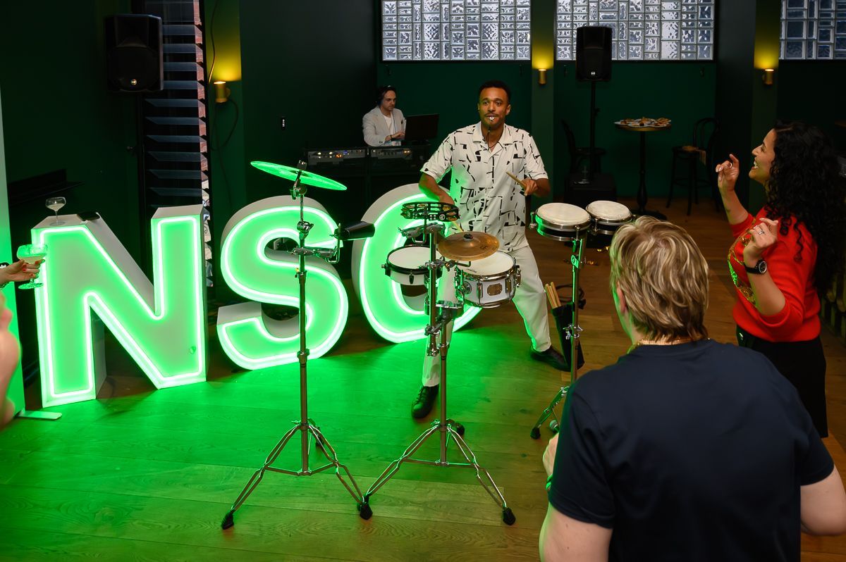 A musician plays small drums in a room lit by large, green glowing letters, while two people dance nearby.