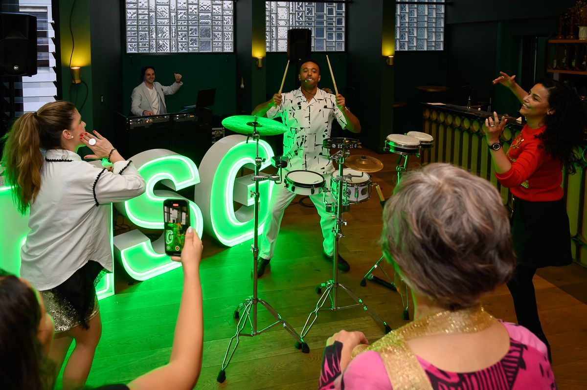 A drummer performs on stage with lit-up neon letters in the background as people dance at an indoor event.