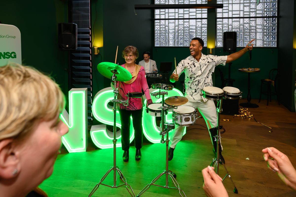 A person plays drums alongside an older individual in front of large, glowing green letters in a dimly lit event space.