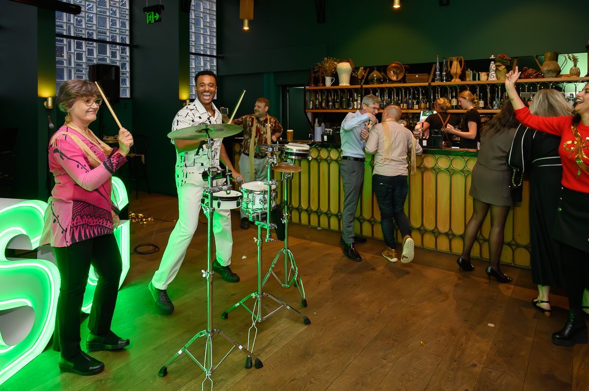 Two people play a drum kit in a bar, while others gather and socialize near the counter in the background.