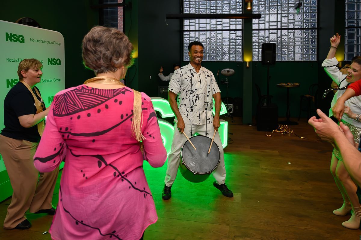 A person in a patterned shirt plays a drum while people dance in a brightly lit room with a green wall backdrop.