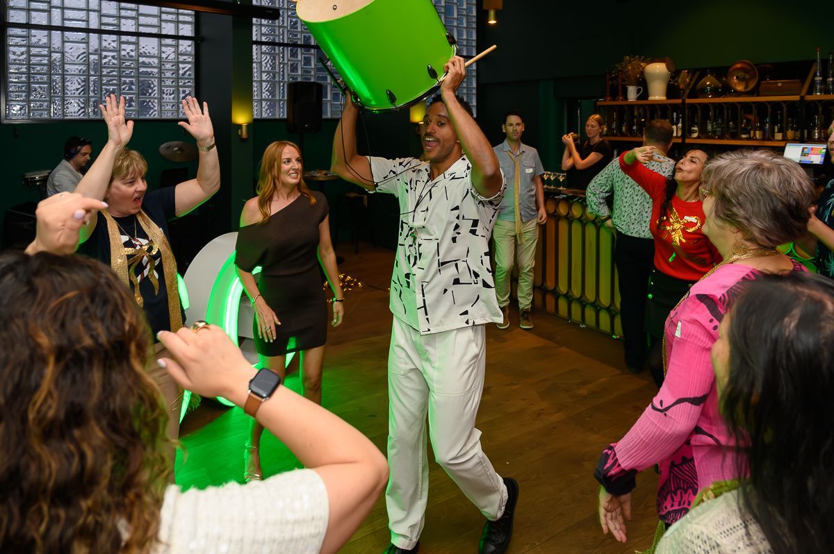 A person lifts a bright green drum overhead while others dance at a social gathering in a dimly lit, festive room.