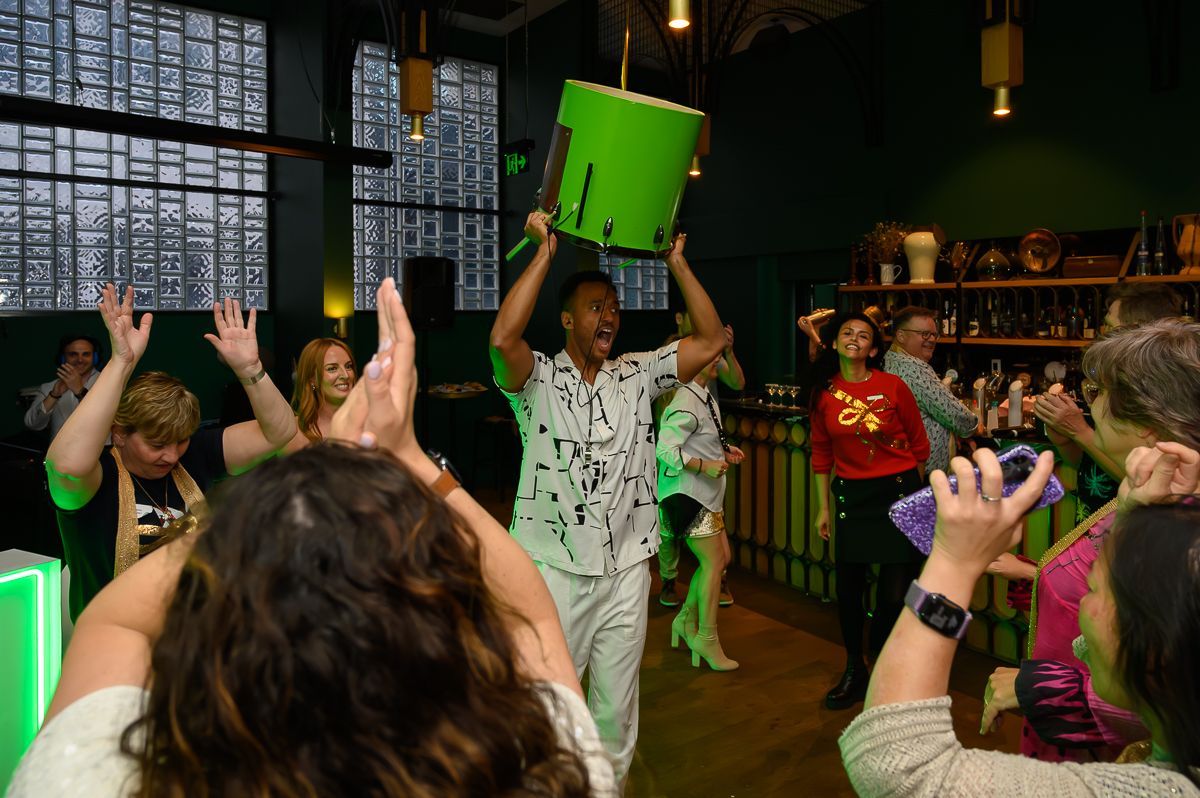 A person in a patterned shirt lifts a lime-green drum over their head while others dance and cheer in a dimly lit venue.