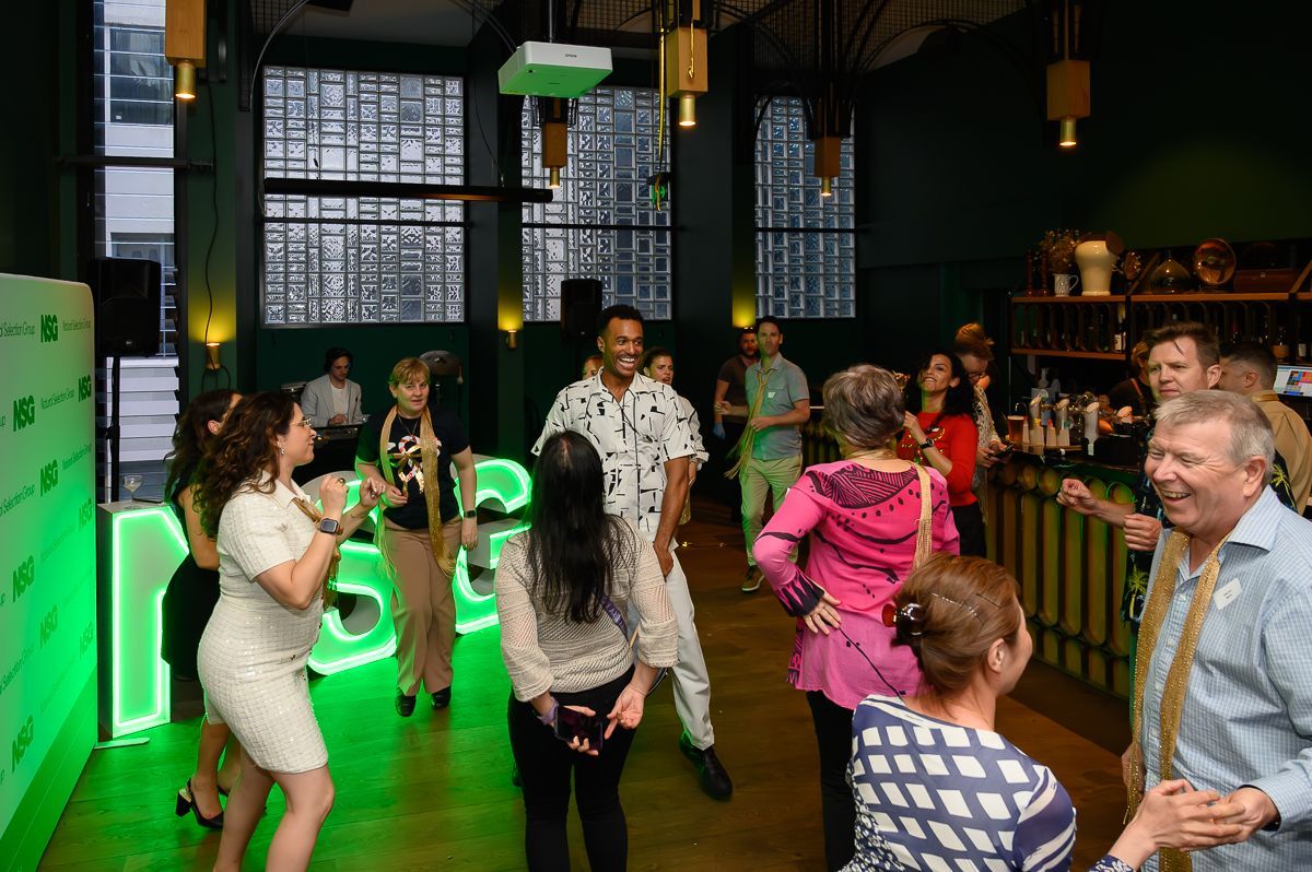 A group of people dancing and smiling in a dimly lit, contemporary lounge space with green accent lighting.