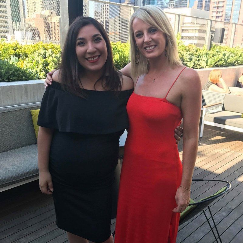 Two people smiling on an outdoor rooftop patio; one wears an off-the-shoulder black dress, the other a red maxi dress.