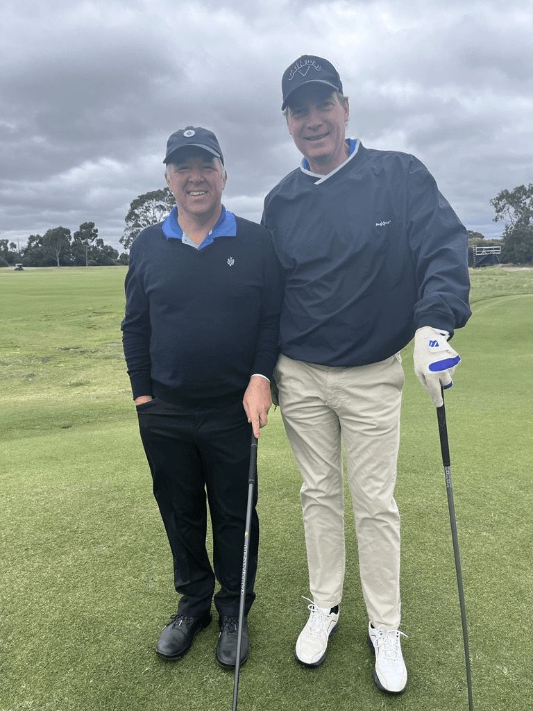 Two men in golf attire and caps smiling while standing on a grassy golf course, both holding golf clubs.